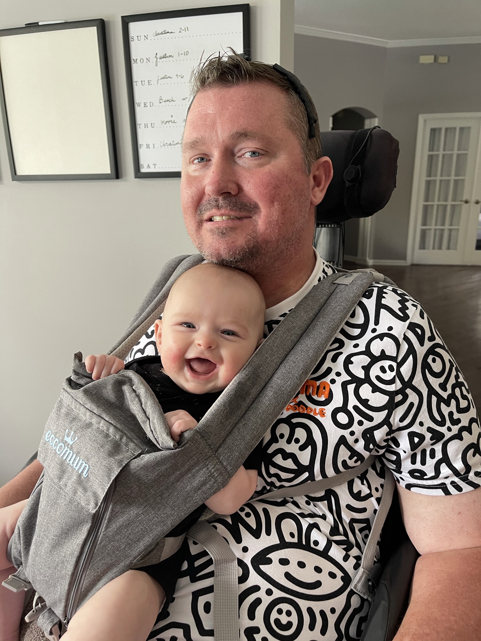Charlie holding a smiling baby in a carrier while seated in his power wheelchair.